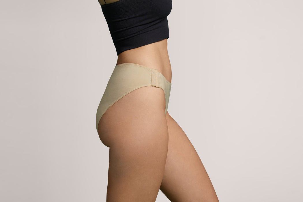 Side angle of a model wearing Slick Chicks Adaptive High Waist Brief in beige against a plain light beige studio background