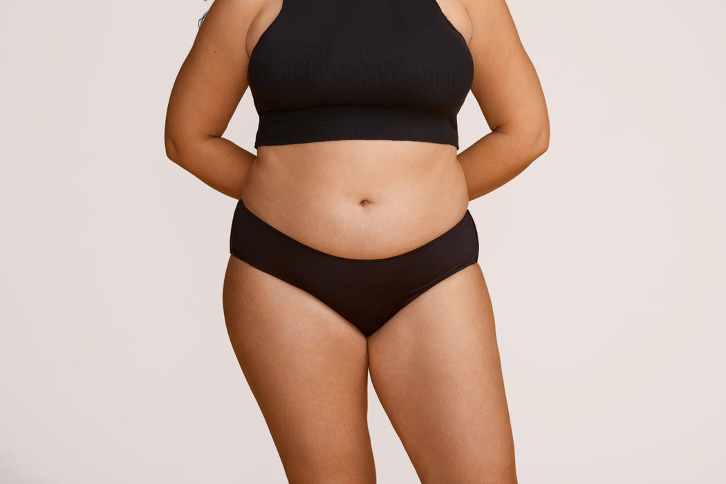 Front angle of a model wearing Slick Chicks Adaptive Hipster Panty in black against a plain light beige studio background