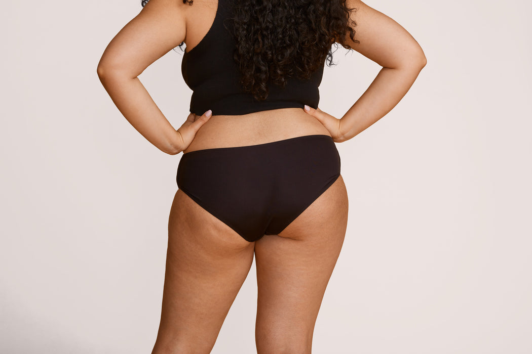 Back angle of a model wearing Slick Chicks Adaptive Hipster Panty in black against a plain light beige studio background