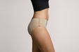 Side angle of a model wearing Slick Chicks Adaptive Hipster Panty in beige against a plain light beige studio background