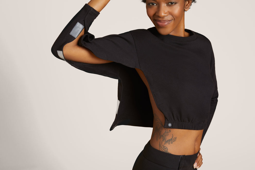 Front angle of woman with short curly black hair wearing Slick Chicks Adaptive Long Sleeve Lounge Shirt in black, with the VELCRO Brand Fasteners open along the side of the Shirt, against a solid beige background