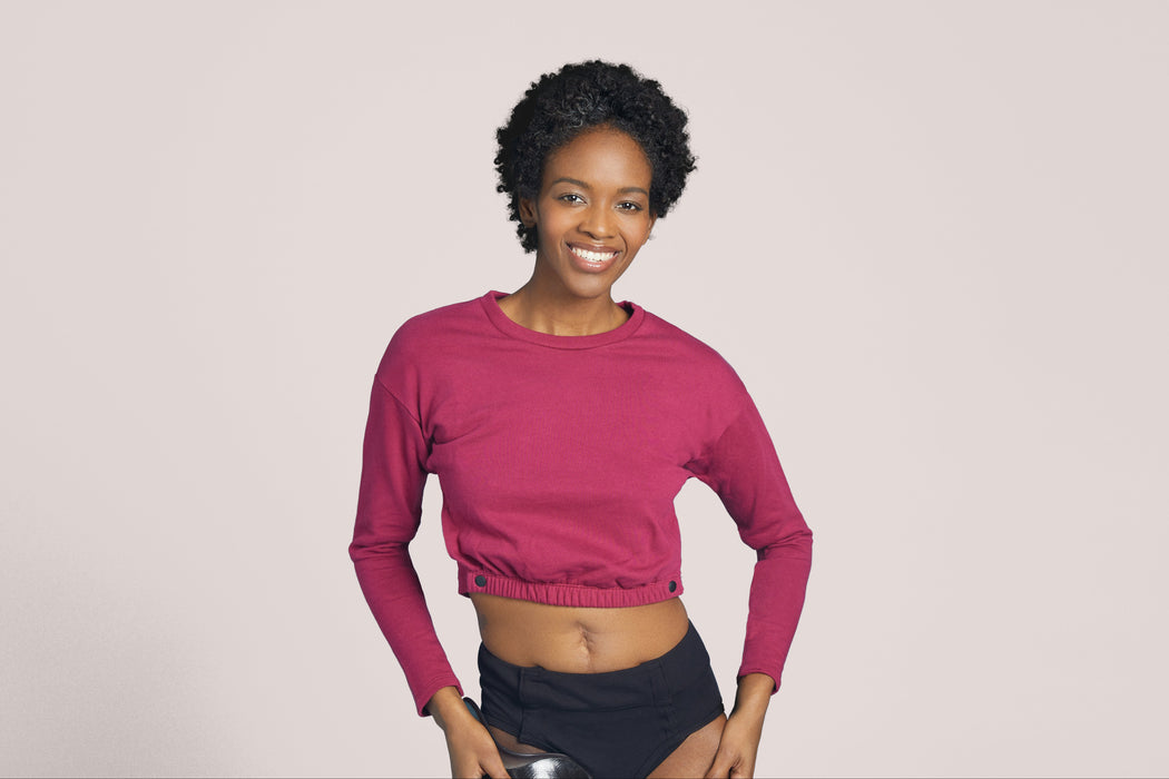 Front angle of a smiling woman with short curly black hair wearing Slick Chicks Adaptive Long Sleeve Lounge Shirt in plum, against a solid beige background
