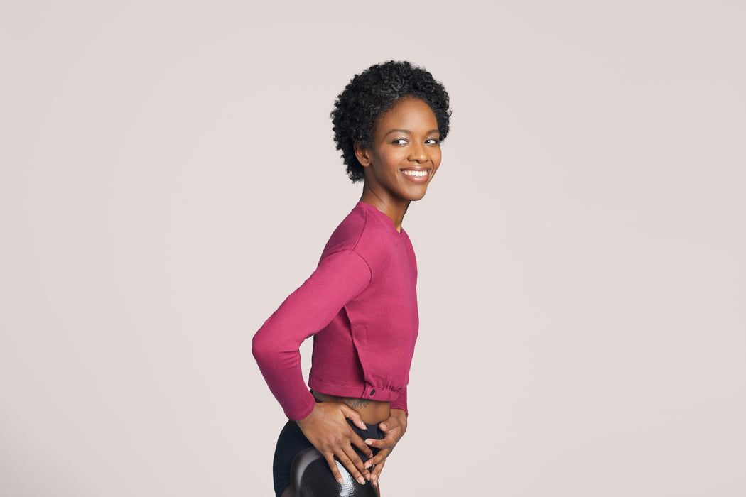 Side angle of a smiling woman with short curly black hair looking to the side wearing Slick Chicks Adaptive Long Sleeve Lounge Shirt in plum, against a solid beige background