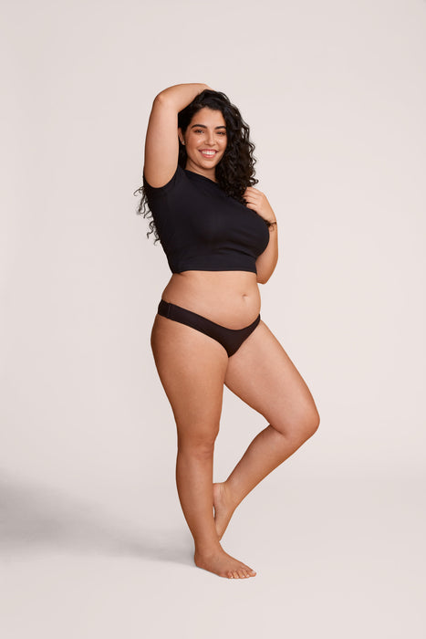 A woman modeling the Slick Chicks Adaptive Tanga Panty in Black, posed against a solid light beige studio background.