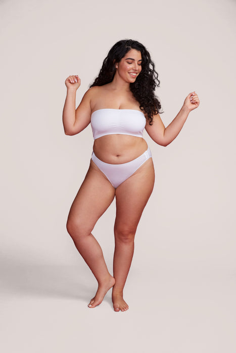 A woman modeling the Slick Chicks Adaptive Tanga Panty in White, posed against a solid light beige studio background.