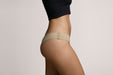 Side view of a model wearing Slick Chicks Adaptive Tanga Panty in Beige against a beige studio background.