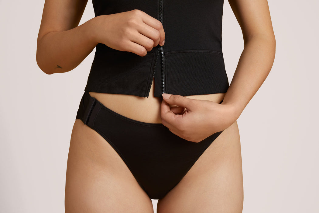 A close-up of a model wearing Slick Chicks Adaptive Two-Way Front Zip Cami in black, demonstrating the two-way front zipper closure, against a plain beige background