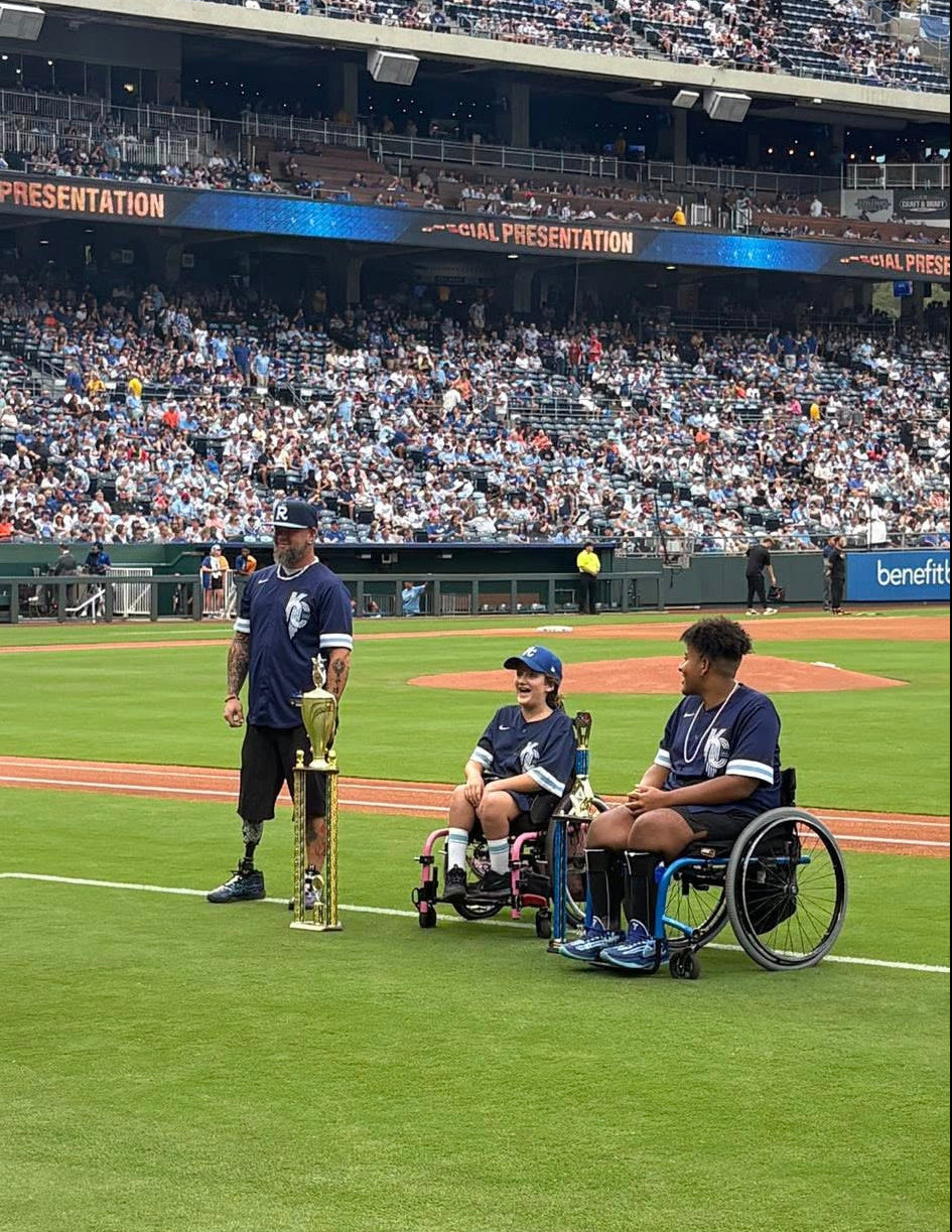 Team Amplife® Ambassador Jason Loftiss with two wheelchair softball athletes from the team he coaches on the Kansas City Royals baseball field with their championship in front of a crowd