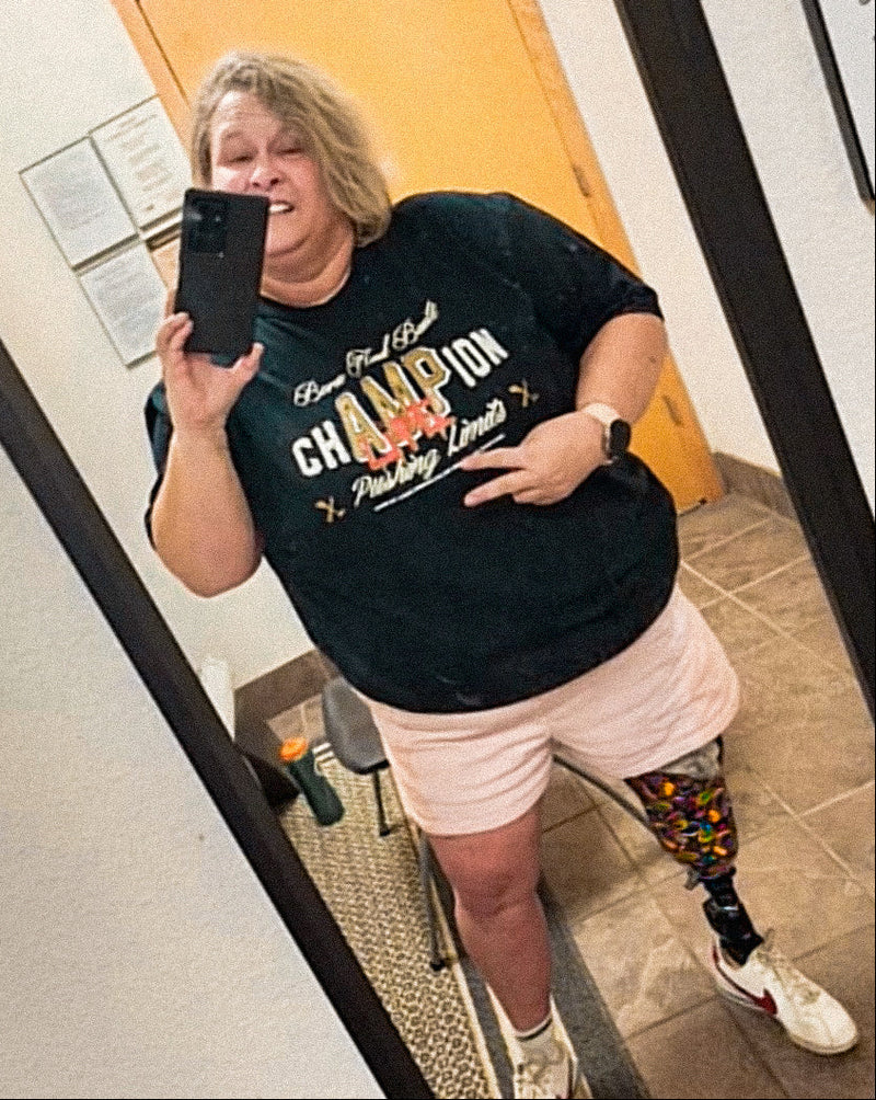 Mirror selfie of Team Amplife® Ambassador Tracy Metzger wearing the Born and Built Champion T-Shirt in her prosthetist office