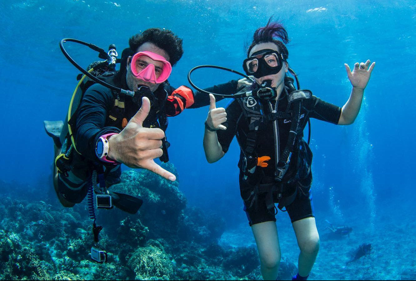 Two scuba divers underwater giving shaka signs with a clear blue ocean background.