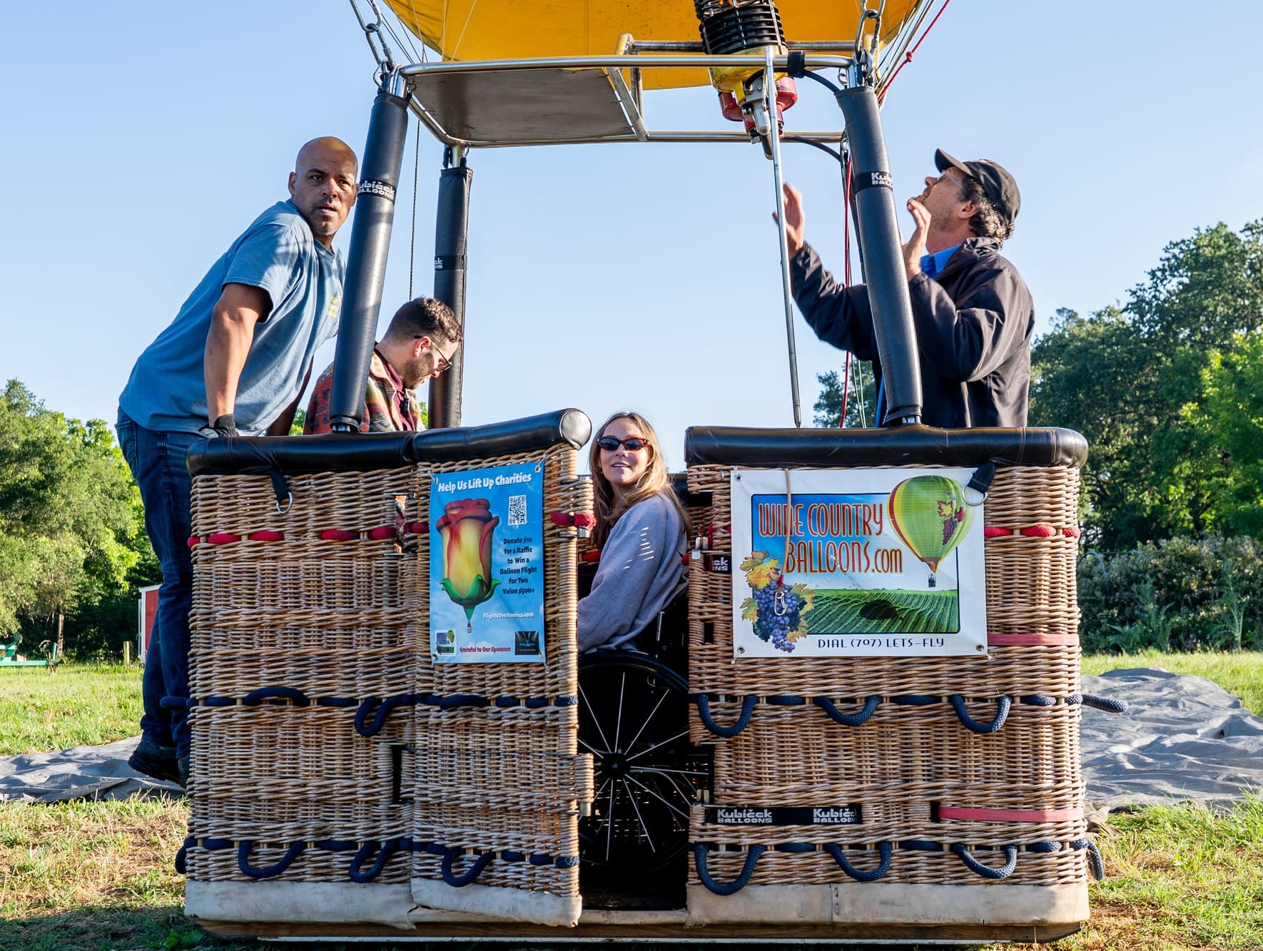 A group of people standing in and around a wicker hot air balloon basket outdoors on a grassy field under a blue sky. They are helping a person using a wheelchair and their partner get setup. The basket has printed signs and a yellow canopy overhead.