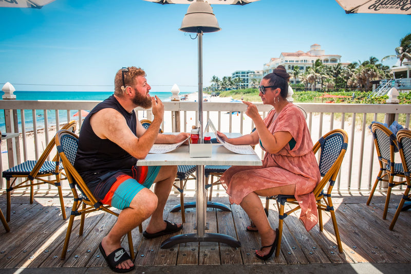 Two people, one with hearing loss, sitting at a table on a beachside deck with ocean view