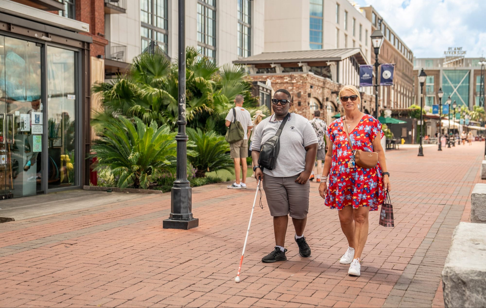 Two people walking on a city street, one with a walking stick, in an urban setting.