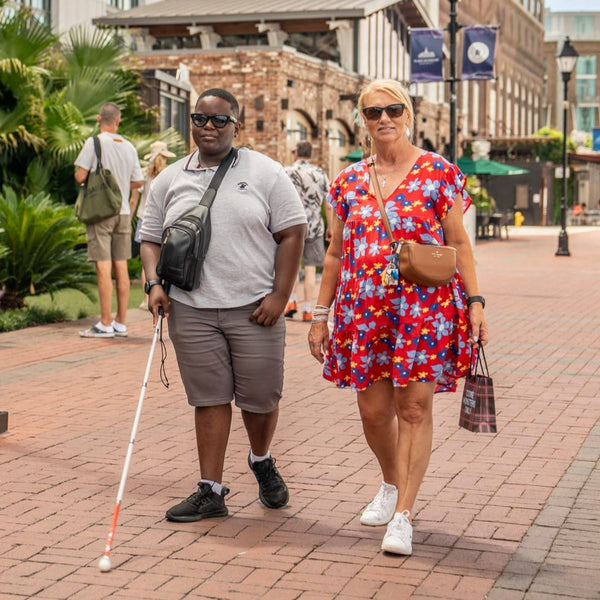 Two people walking on a city street, one with a walking stick, in an urban setting.
