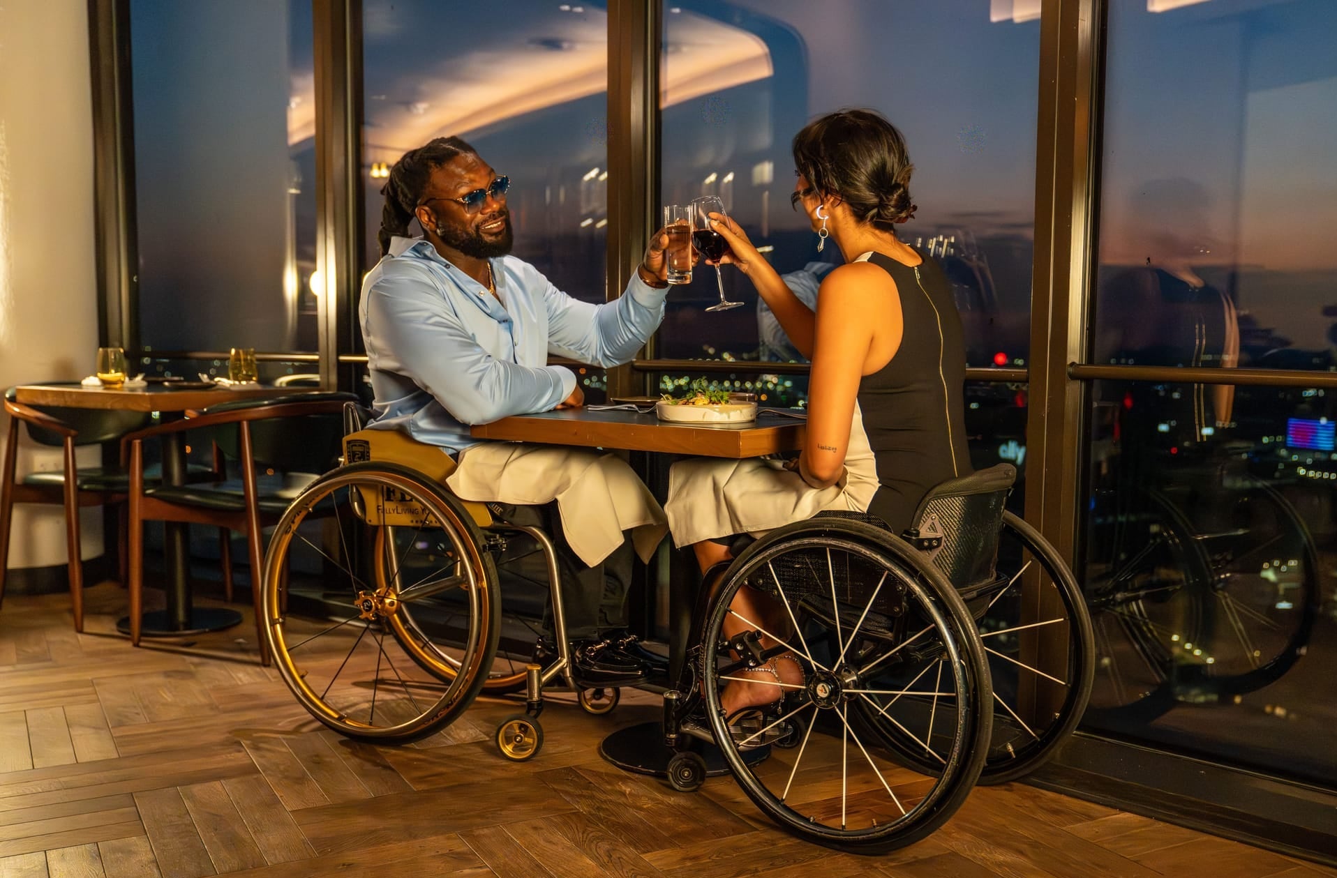 Two people using wheelchairs enjoying a romantic evening at a high-rise restaurant with city lights in the background.