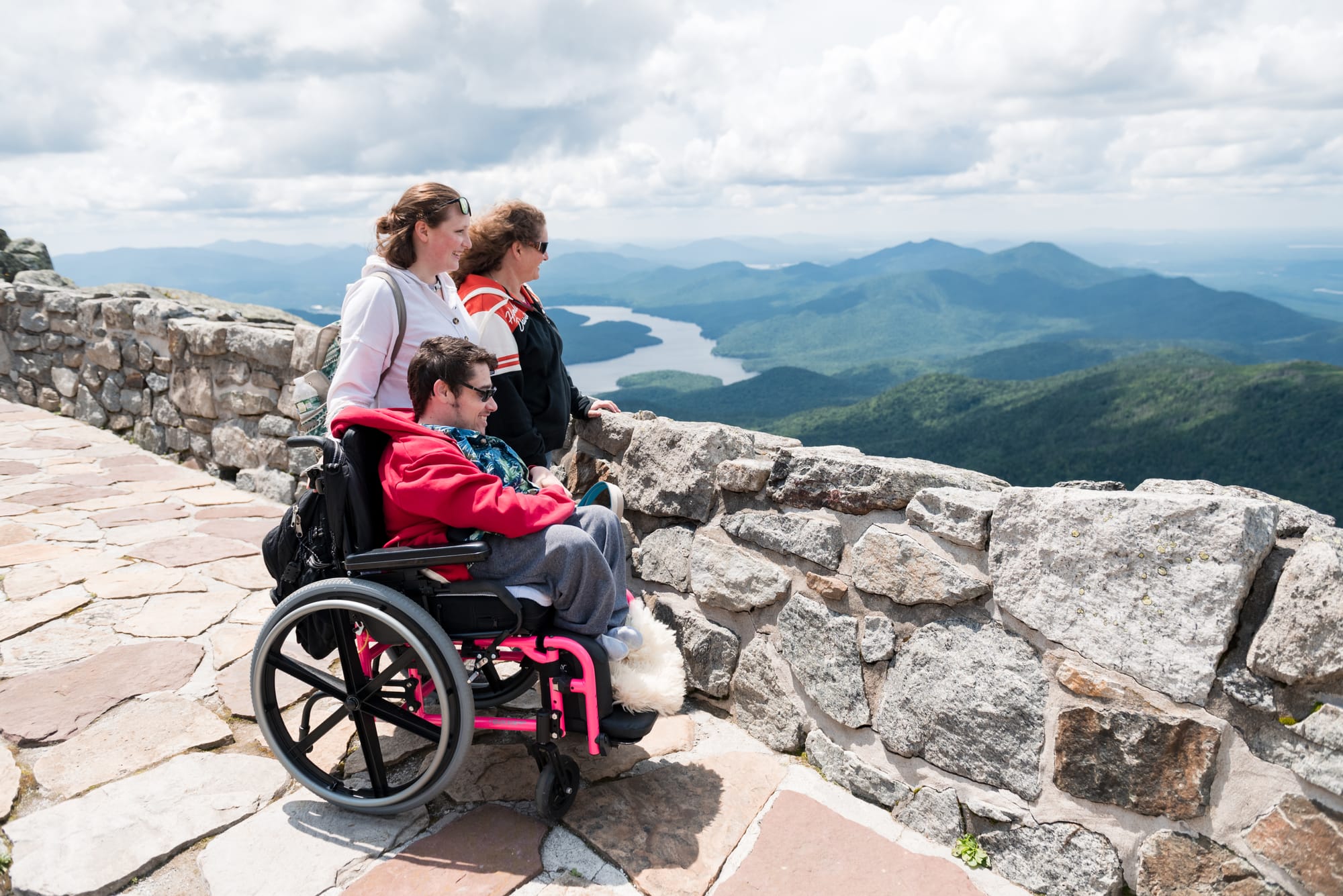 Person using a wheelchair with two others overlooking a scenic mountain view.