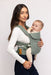 Side of woman holding a baby in a WildBird Acadian Aerial Buckle Baby Carrier on a white background