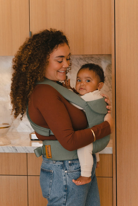 Woman holding a baby in a WildBird Acadian Aerial Buckle Baby Carrier in a kitchen setting