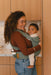 Woman holding a baby in a WildBird Acadian Aerial Buckle Baby Carrier in a kitchen setting