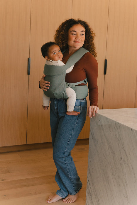 Woman holding a baby in a WildBird Acadian Aerial Buckle Baby Carrier in a kitchen with wooden cabinets