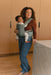 Woman holding a baby in a WildBird Acadian Aerial Buckle Baby Carrier in a kitchen with wooden cabinets