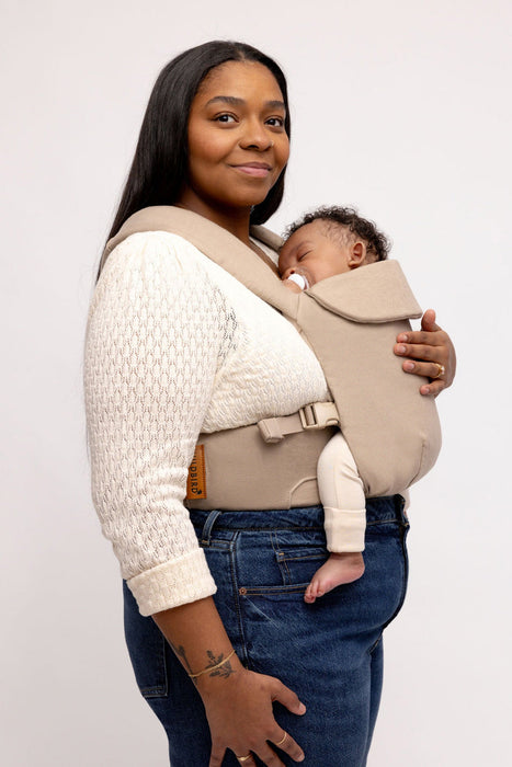 Side of woman holding a baby in a WildBird Desert Lark Aerial Buckle Baby Carrier on a white background