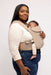 Side of woman holding a baby in a WildBird Desert Lark Aerial Buckle Baby Carrier on a white background