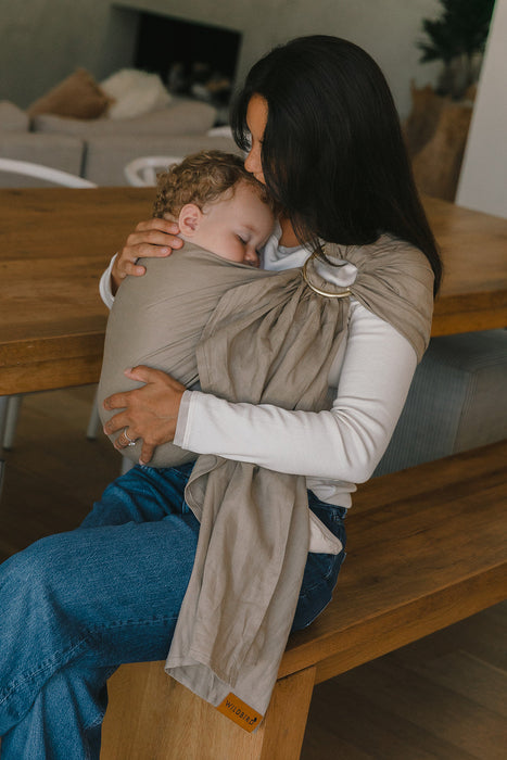 Woman sitting on a wooden bench holding and kissing a baby in a WildBird Desert Lark Linen Baby Sling inside a home.