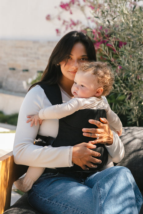 Woman holding a baby in a WildBird Raven Aerial Buckle Baby Carrier outdoors with plants in the background
