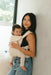 Woman holding a baby in a WildBird Sparrow Aerial Buckle Baby Carrier against a light-colored wall.