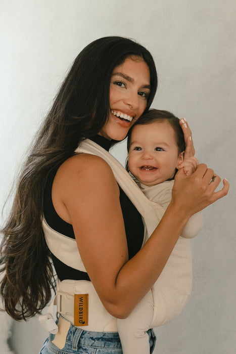 Woman holding a baby in a WildBird Sparrow Aerial Buckle Baby Carrier  against a plain background