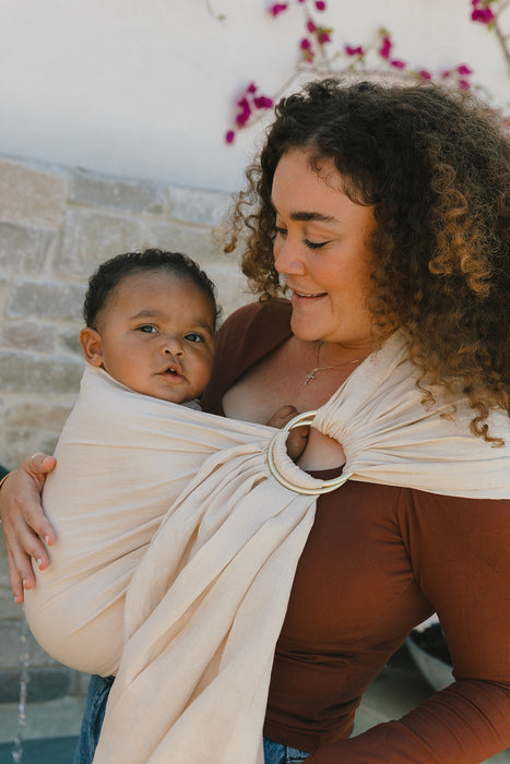 Woman holding a baby in a WildBird Sparrow Linen Baby Sling against a stone wall and floral background