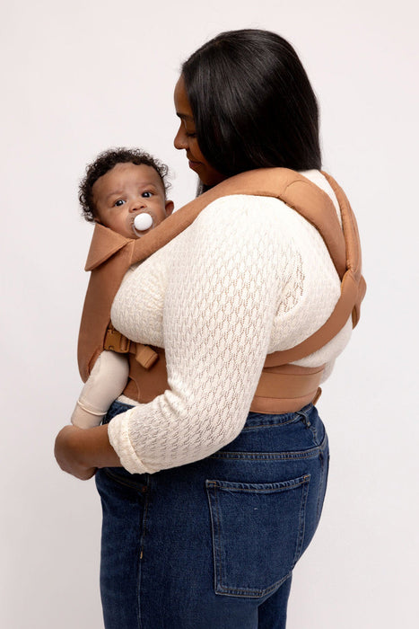 Side of woman holding a baby in a WildBird Willow Aerial Buckle Baby Carrier on a white background