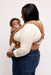 Side of woman holding a baby in a WildBird Willow Aerial Buckle Baby Carrier on a white background
