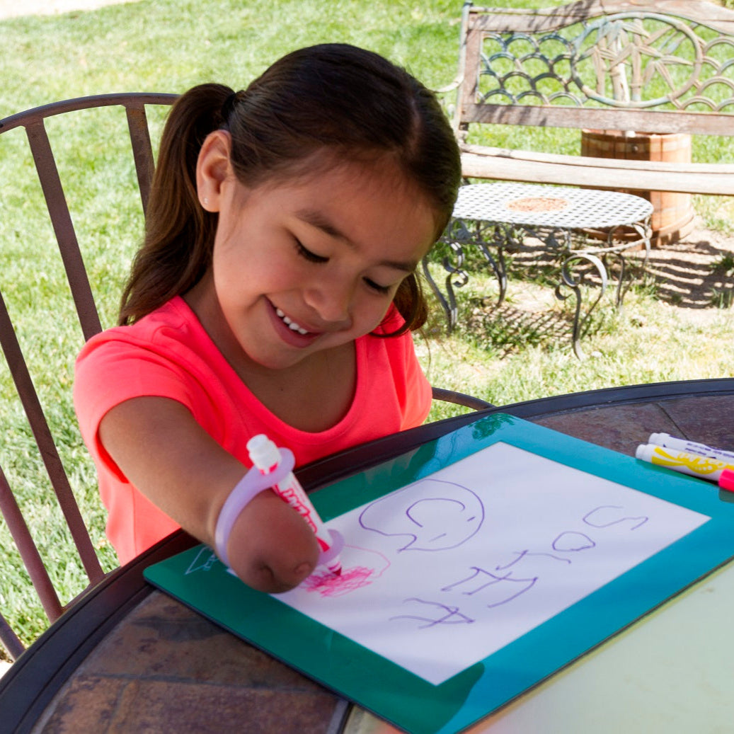 Young girl drawing on paper using the EaZyHold with markers at a table outdoors.