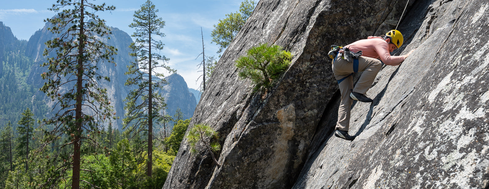 Person climbing a rocky mountain face with trees and mountains in the background