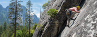 Person climbing a rocky mountain face with trees and mountains in the background