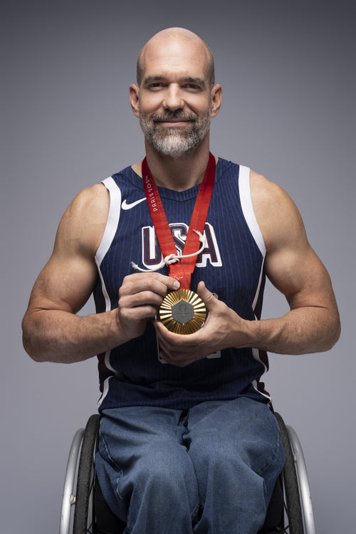 Team USA Wheelchair Basketball player Paul Schulte holding his Paris 2024 gold medal in a Top End wheelchair wearing a USA tank top against a gray background.